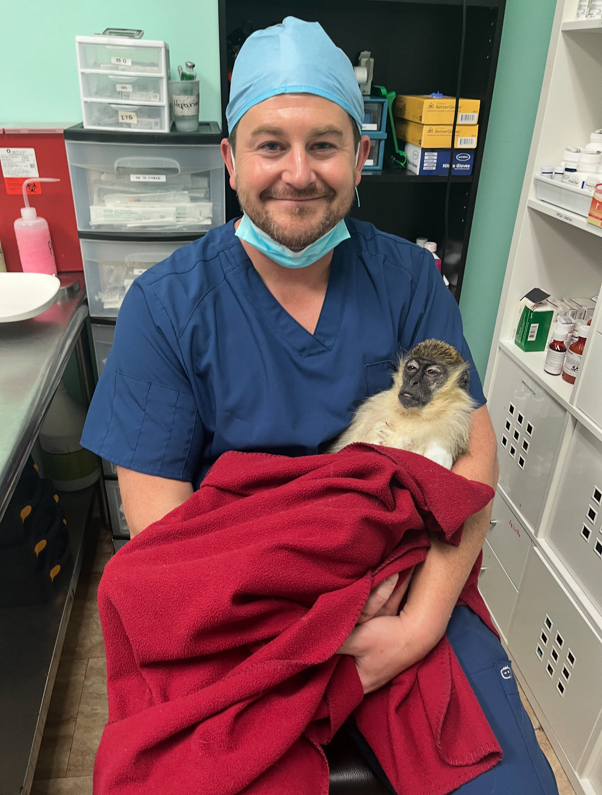 Person in blue scrubs holding a small animal wrapped in a red blanket in a medical setting.